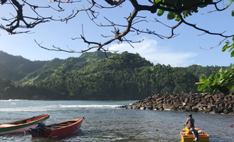 Boats on a calm bay with rocky shore, lush green hills, and a person in a yellow boat under a clear blue sky.