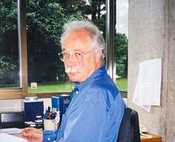 A man sits at his desk in an office. A window looking onto grass and trees is behind him.