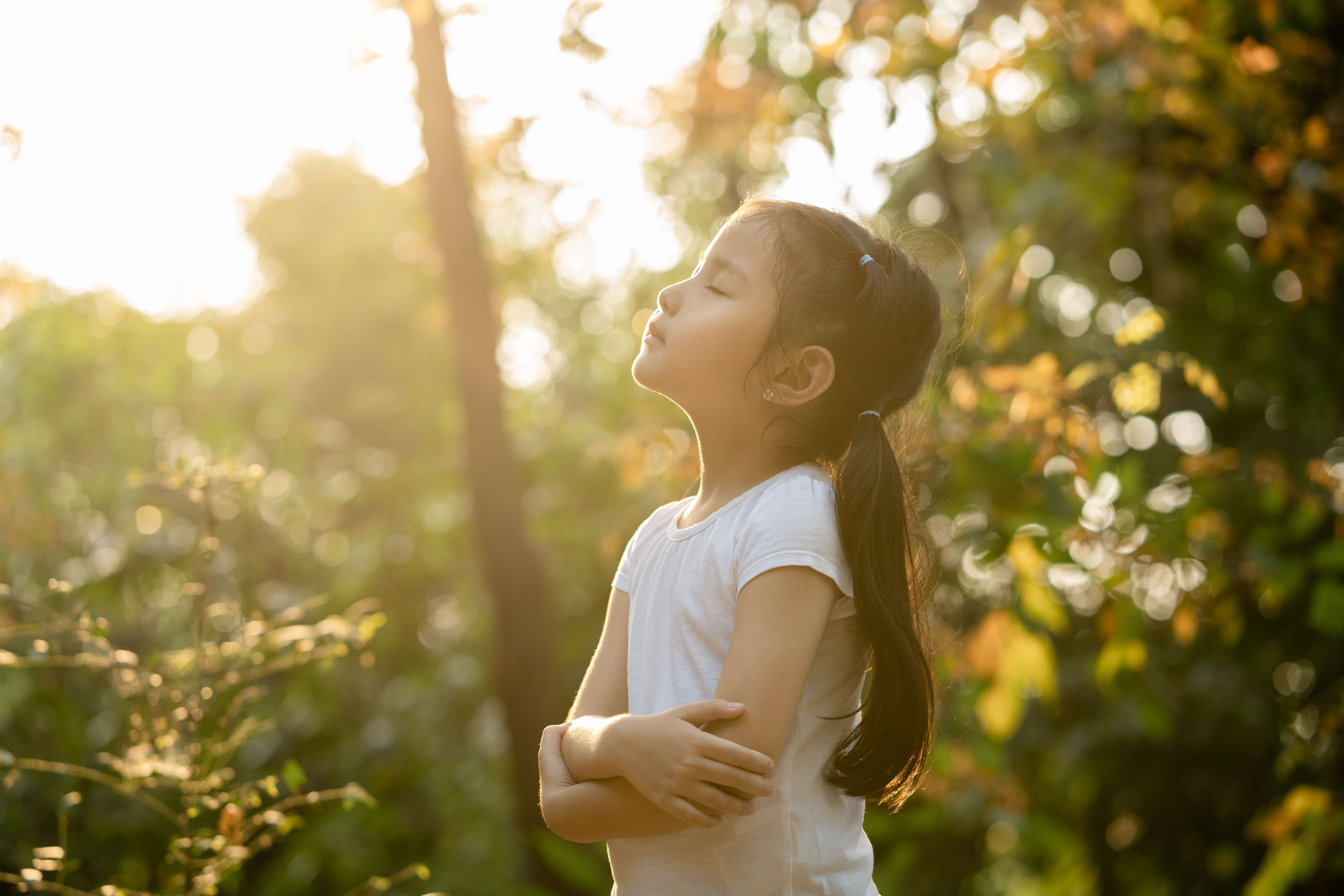Young female child mindfully looking up into the sunshine with closed eyes in the woods