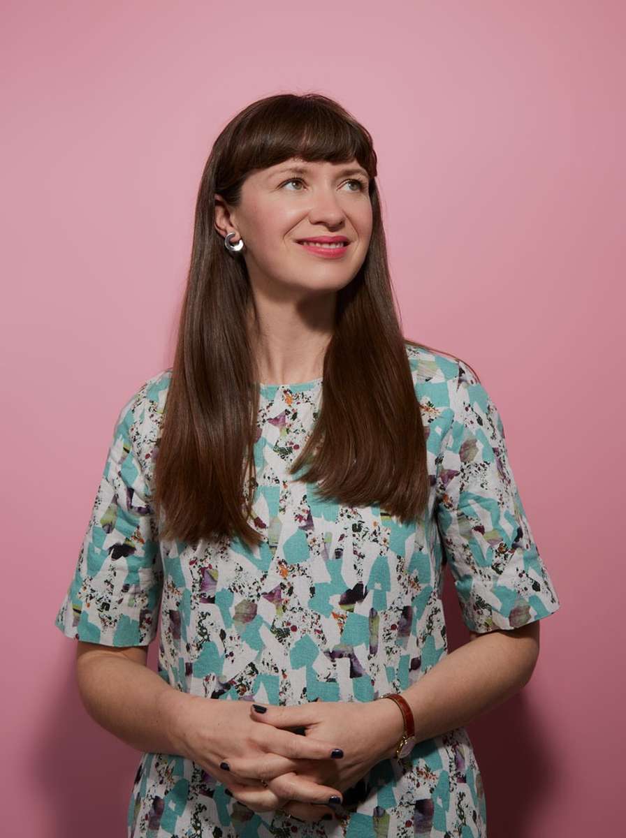 Someone with long brown hair smiles in front of a pink background.