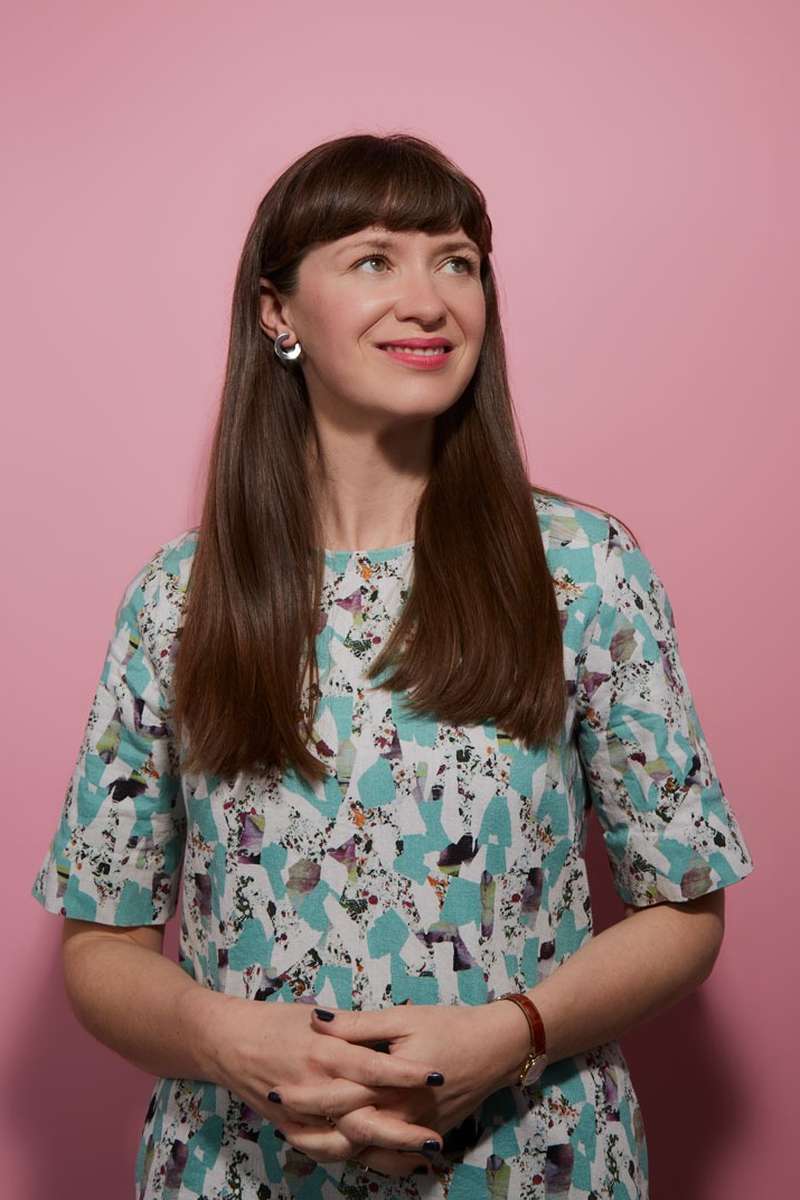 Someone with long brown hair smiles in front of a pink background.