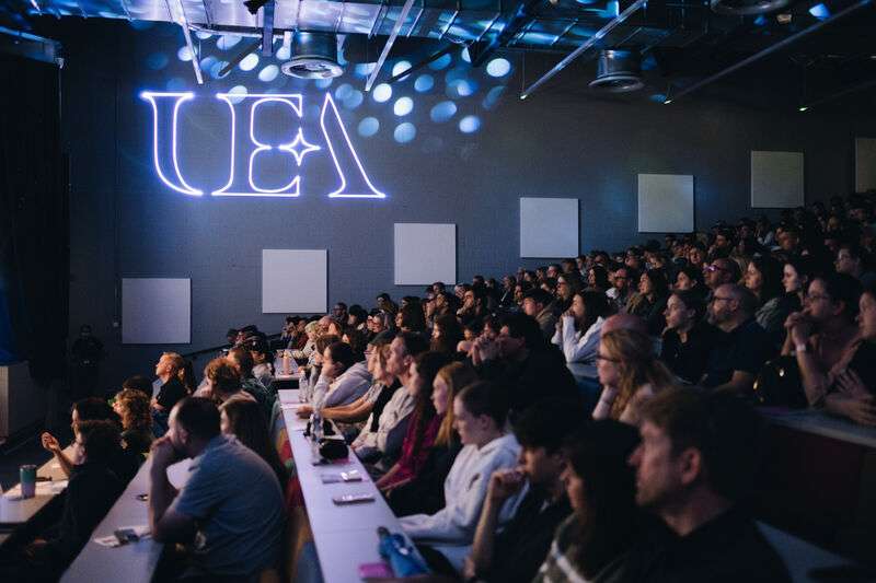 Audience attentively watching a presentation in a dimly lit auditorium, with "UEA" illuminated on the wall.