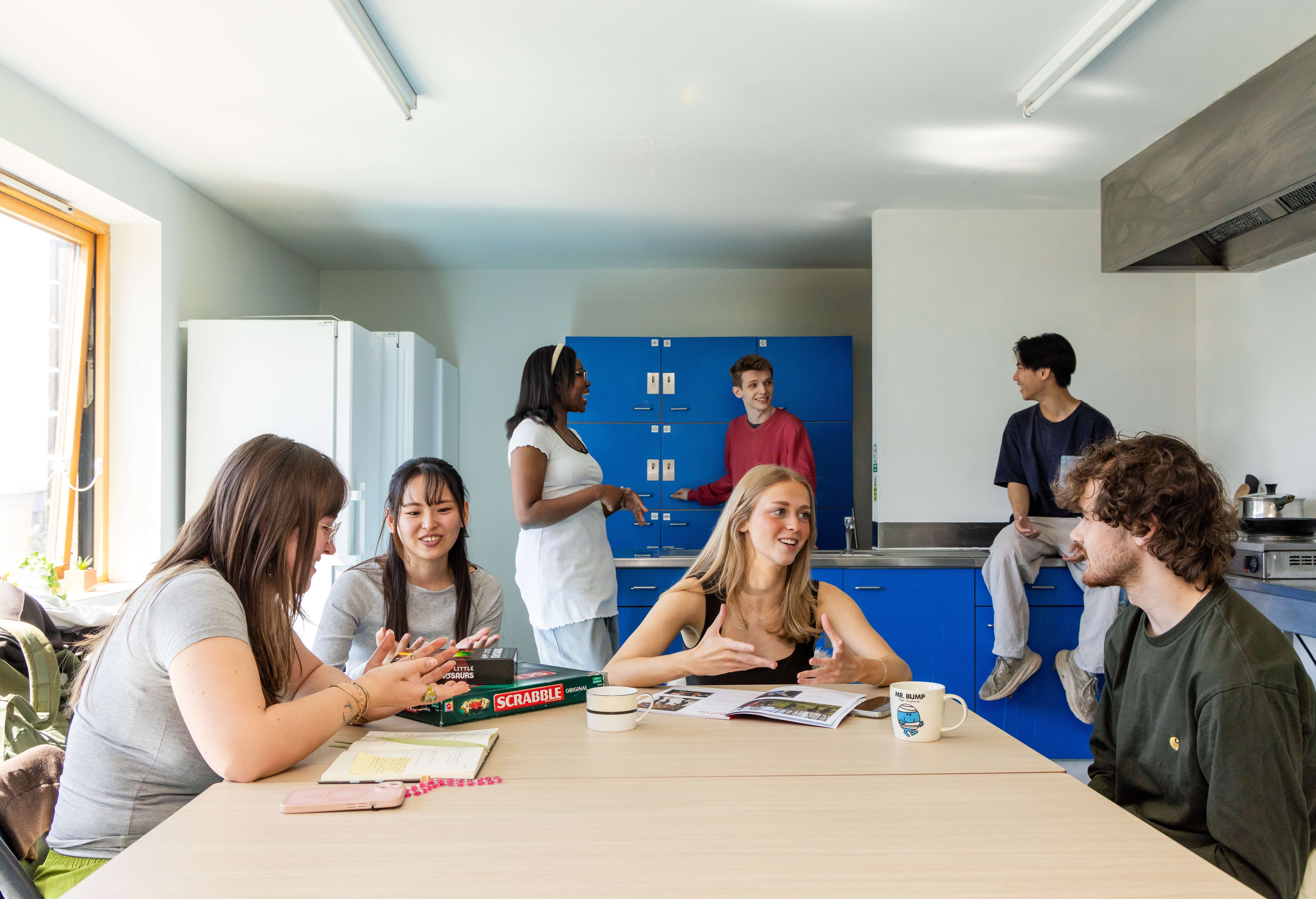 A group of university students laughing and playing board games together in a modern, shared student kitchen.