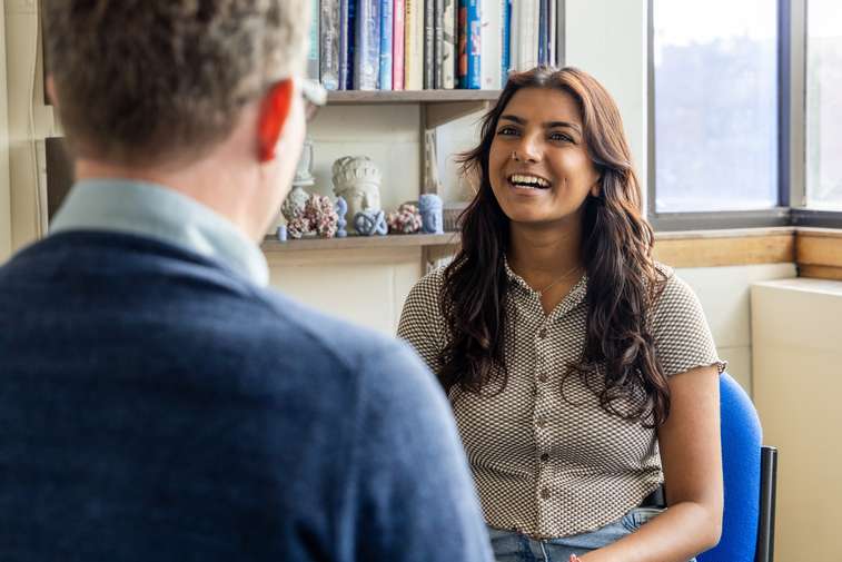 A student has a discussion with a tutor, whose back is to the camera. The student is smiling broadly and looks confident and comfortable