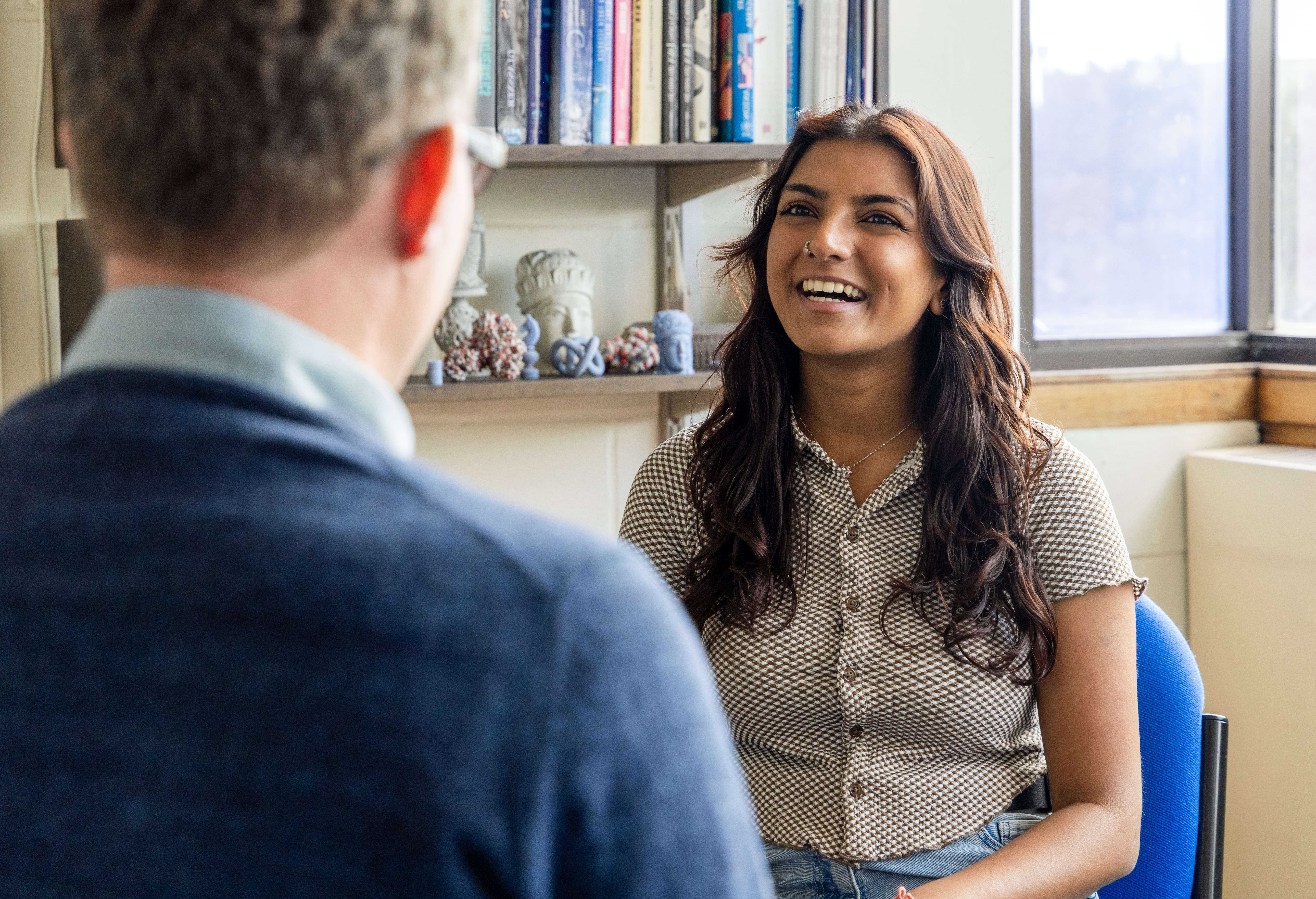 A student has a discussion with a tutor, whose back is to the camera. The student is smiling broadly and looks confident and comfortable