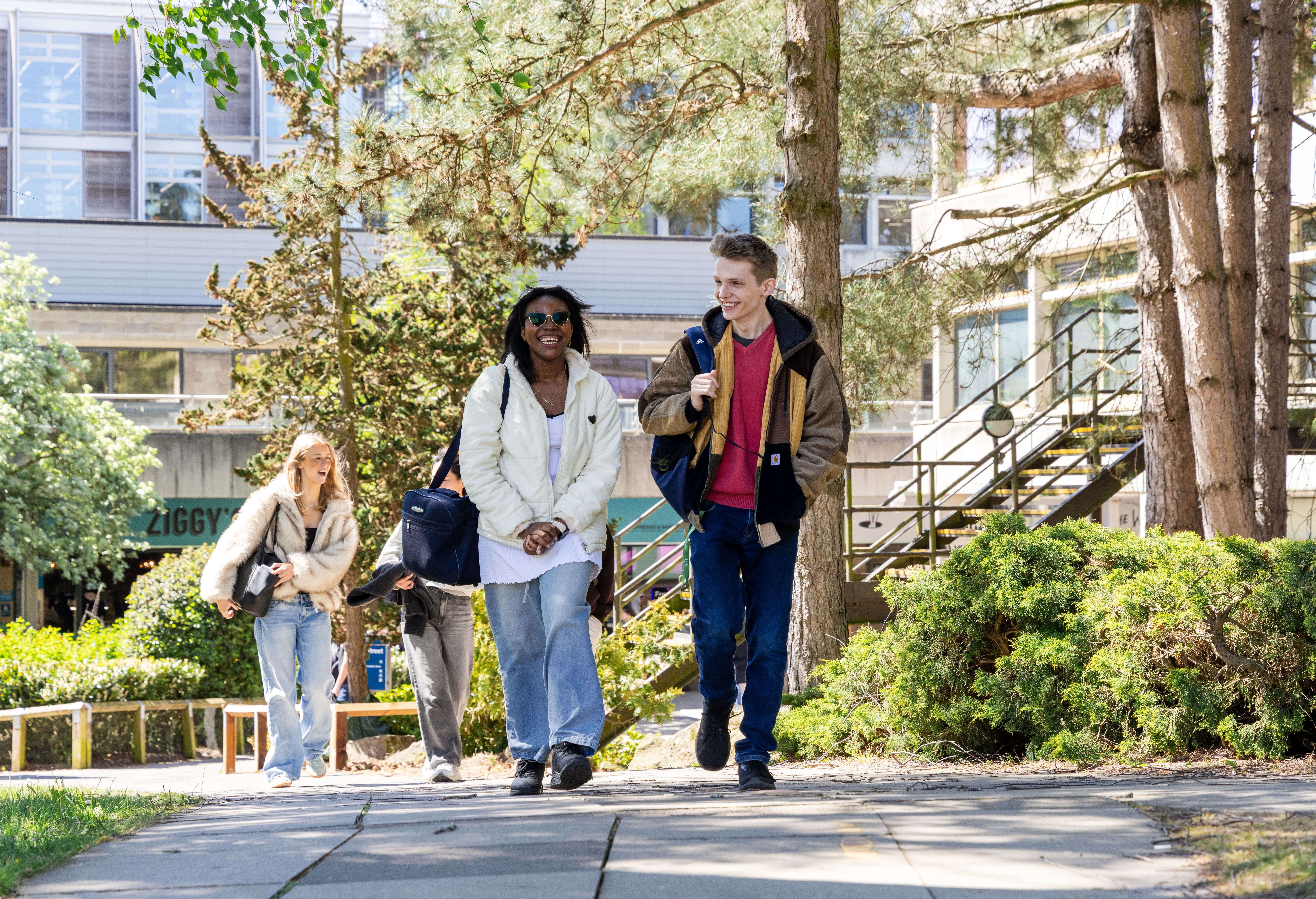 A group of students walks and talks on a paved path through their sunny university campus, with modern buildings and trees behind them.