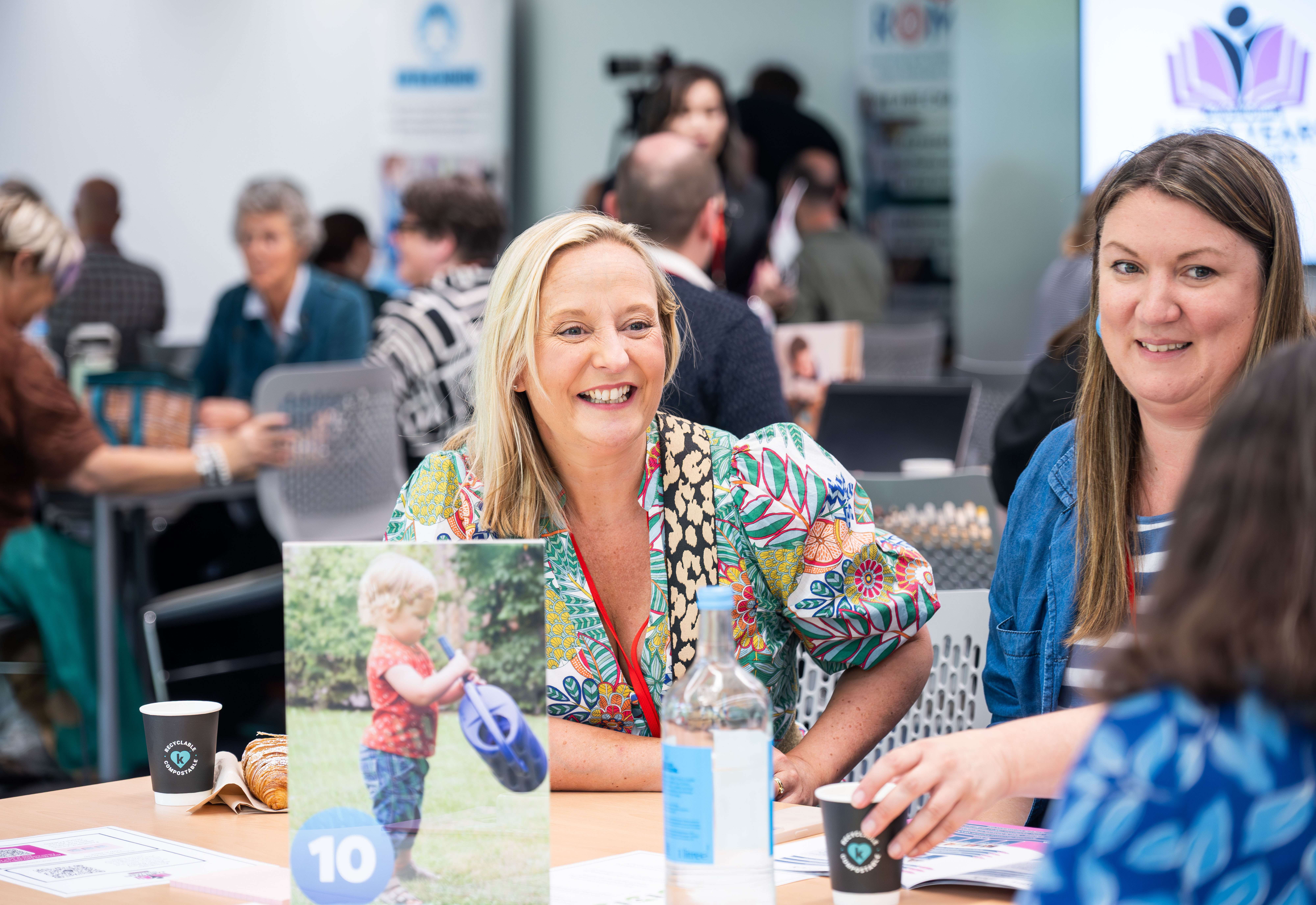 A smiling researcher engages in discussion at a collaborative academic symposium in Norwich, with other attendees networking in the background.