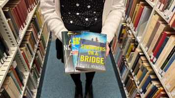 Person in a library aisle holding three books toward camera; front book titled "Strangers on a Bridge" by Louise Mangos.