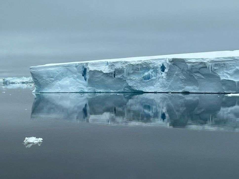 A pale blueish grey iceberg on water so still it looks like glass across a grey background.