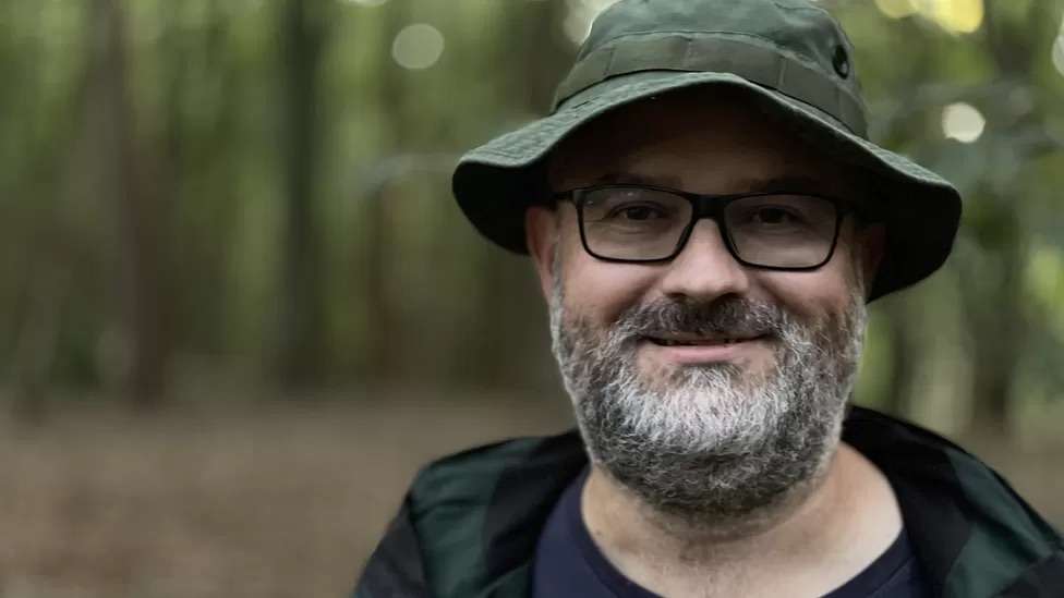 A man in a hat and glasses smiles at the camera. He is in a forest but the background is blurred.